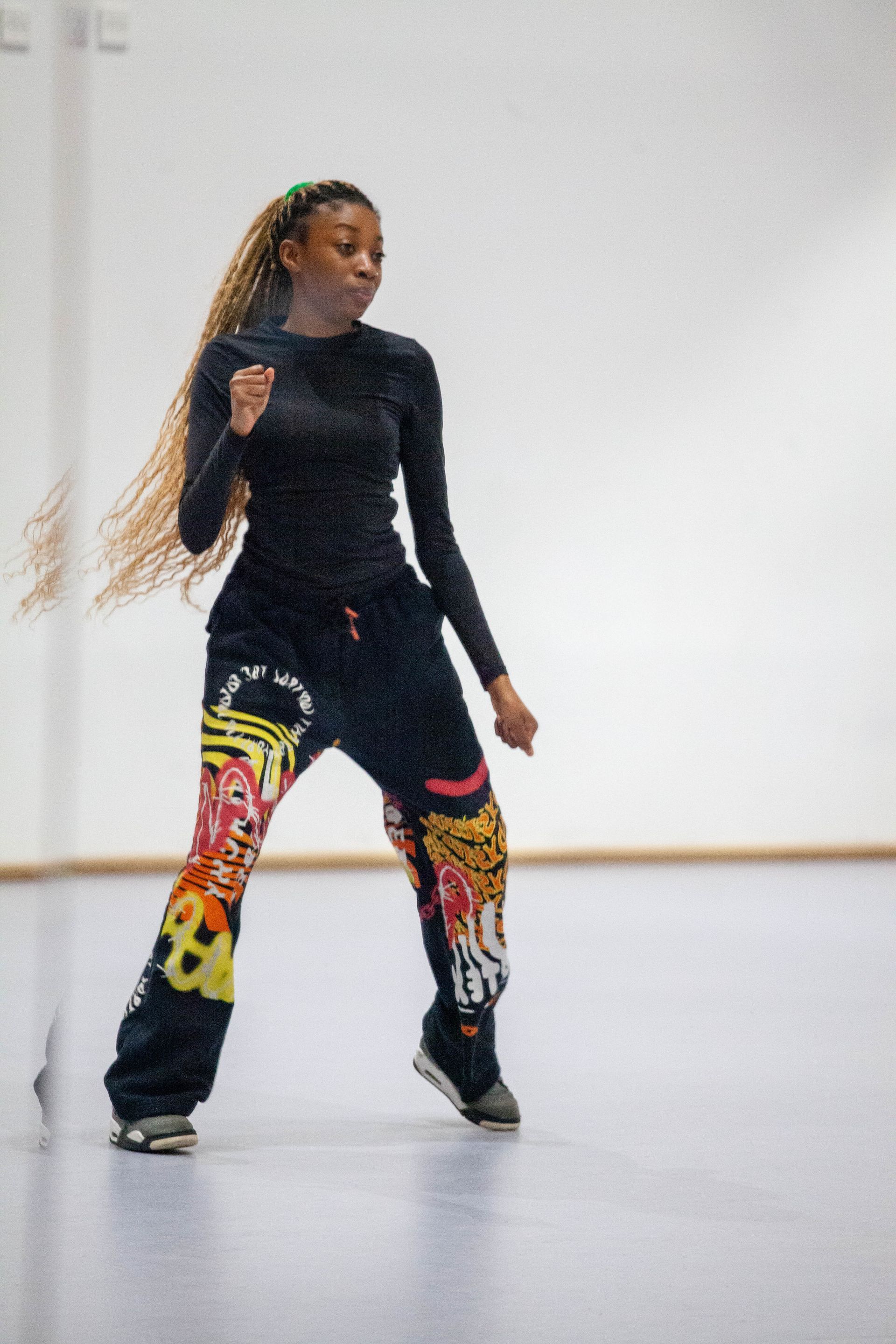 A person with long, light-colored braids stands in a studio, wearing a black long-sleeved shirt and patterned sweatpants.