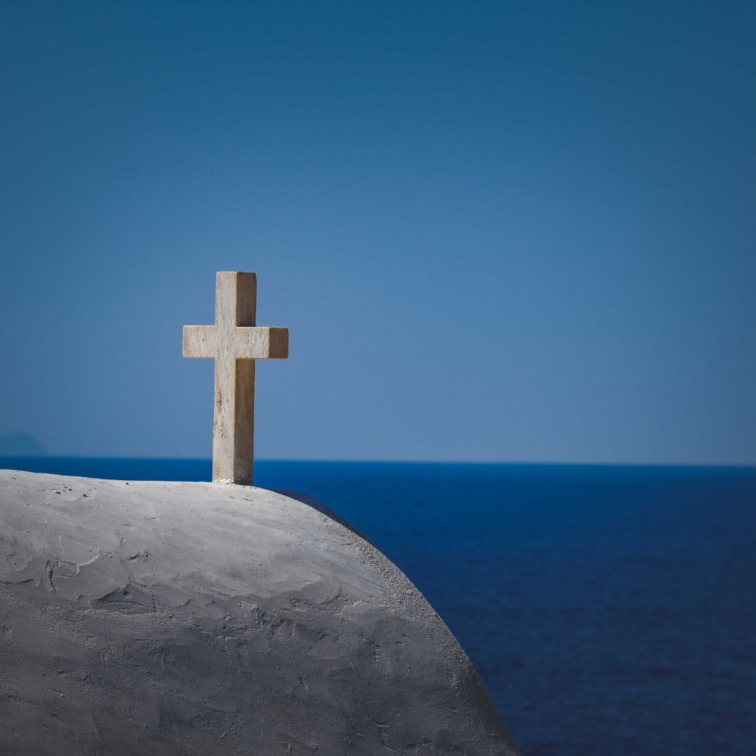 A stone cross stands on a curved, white rooftop overlooking the deep blue sea under a clear sky.