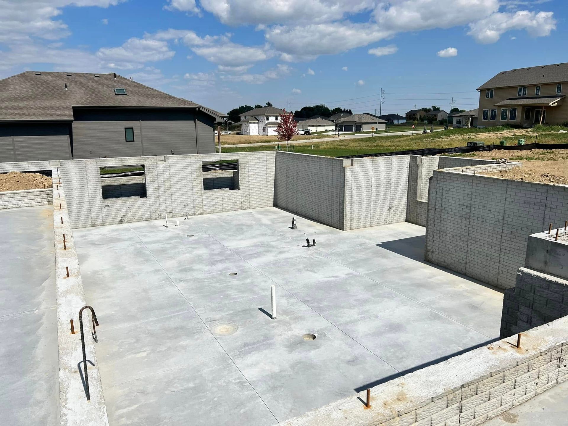 Concrete foundation walls under construction in a suburban neighborhood, with houses and blue sky in the background