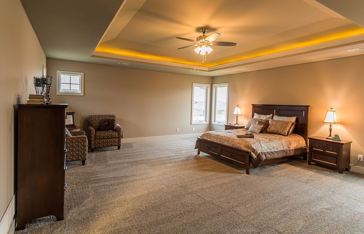 Spacious beige bedroom with a king bed, chairs, nightstands, and a tray ceiling with warm lighting.