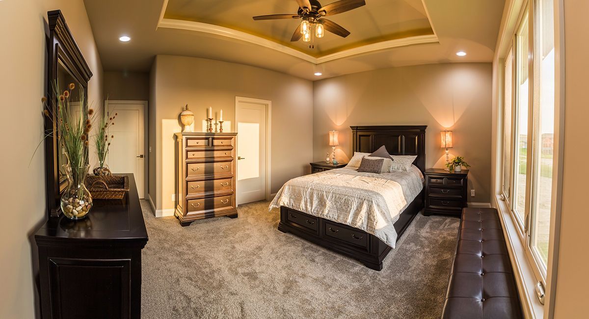 Spacious beige bedroom with a bed, dresser, ceiling fan, and large windows with curtains.