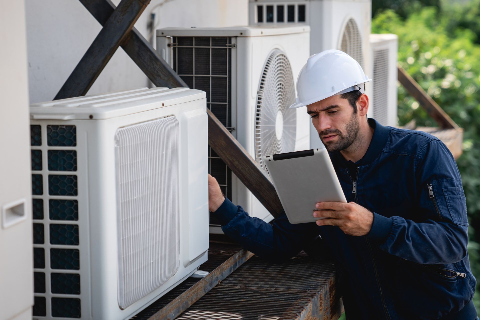 A contractor wearing a helmet and holding a clipboard is inspecting an AC system outdoors.