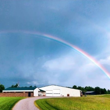 Rainbow arcs over a white building with a green roof, set against a cloudy blue sky. A gravel road leads to it.
