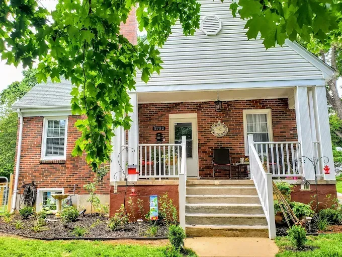 Red brick house with white porch, steps, and trim, framed by green trees and a garden.