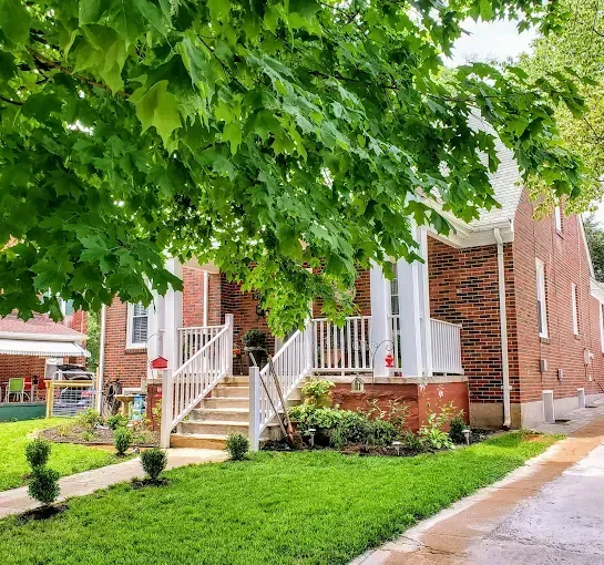 Red brick house with white pillars, porch, and green lawn under a leafy tree.