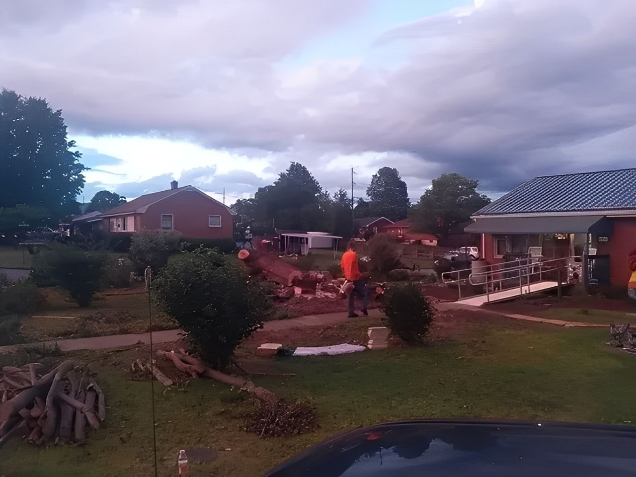 A person working outside in a yard, trees and houses under a cloudy sky.