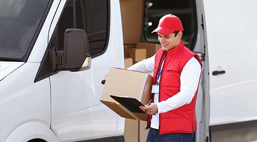 Delivery person in red vest and cap, loading cardboard box into white van.