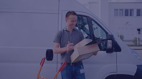 Delivery person smiles, opening a cardboard box next to a white van and a hand truck outdoors.