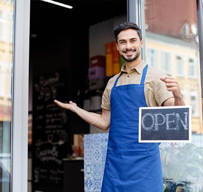 A woman in an apron is holding a tablet in front of a welcome open sign.