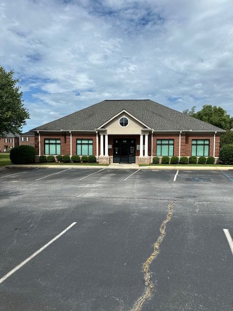 Brick building with dark roof, windows, and a parking lot on a cloudy day.