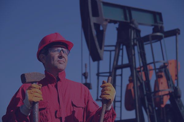 Oil worker in red overalls and hard hat, holding tools, stands before an oil pump.