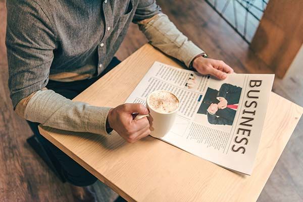 A man is sitting at a table reading a newspaper and drinking coffee.