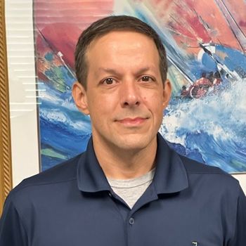 A man in a blue shirt is standing in front of a painting of a boat in the ocean.