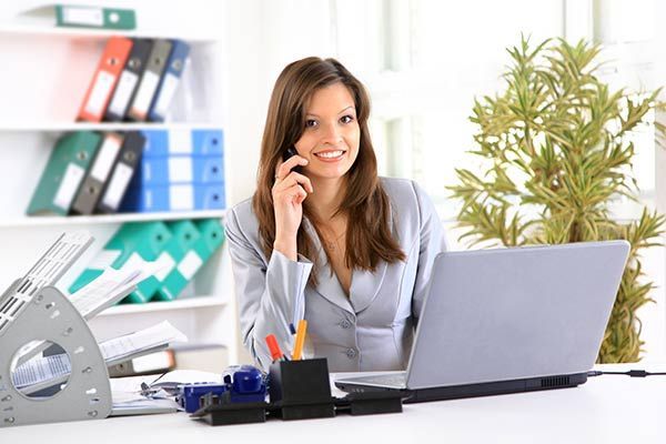 A woman is sitting at a desk with a laptop and talking on a cell phone.