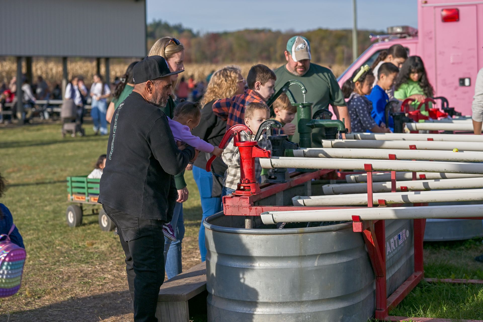 Family at a carnival game: father helps child with a rubber duck race while mother looks on. Outdoors, sunny.