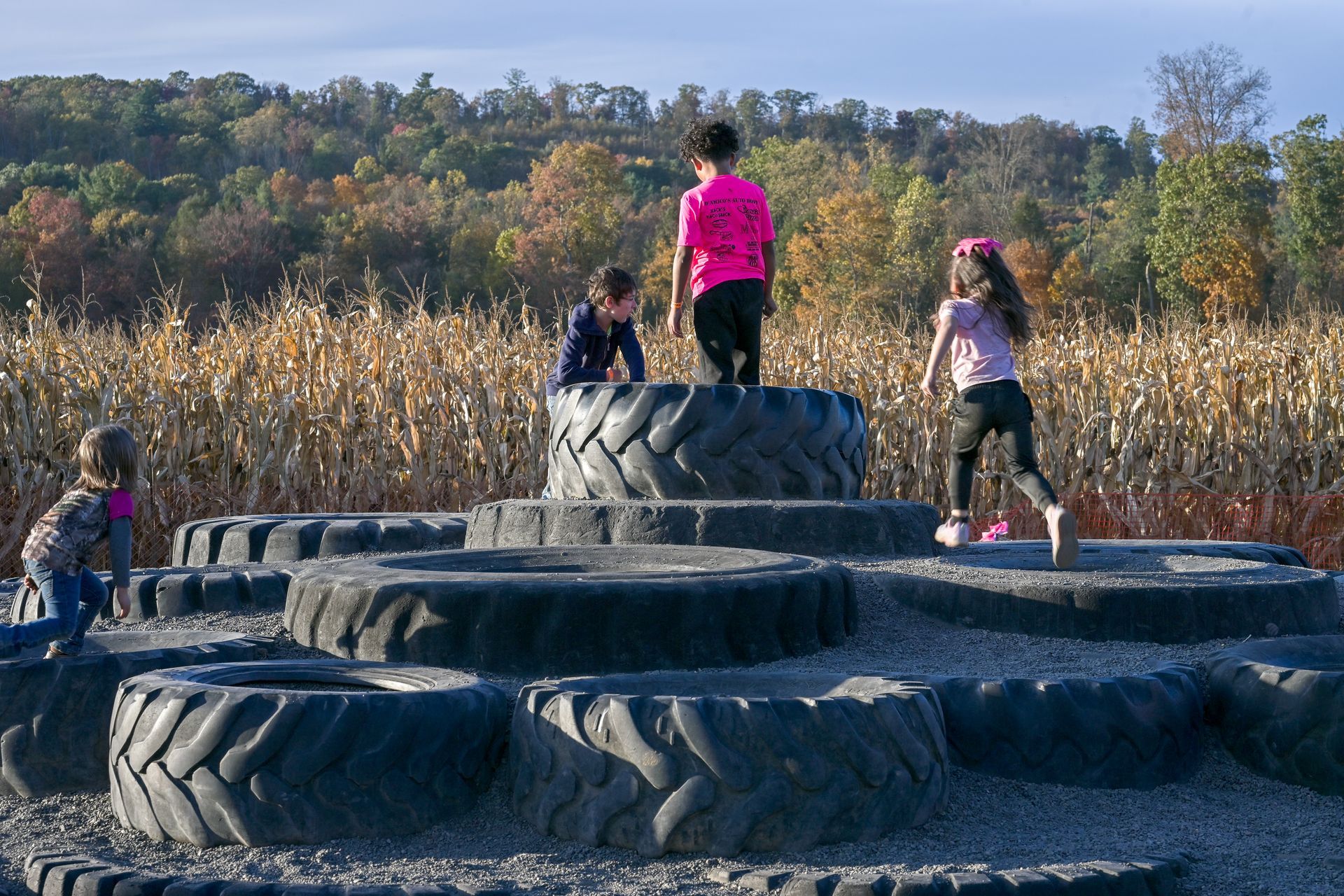 Two children peek over a large tire in a field. Fall foliage in the background.