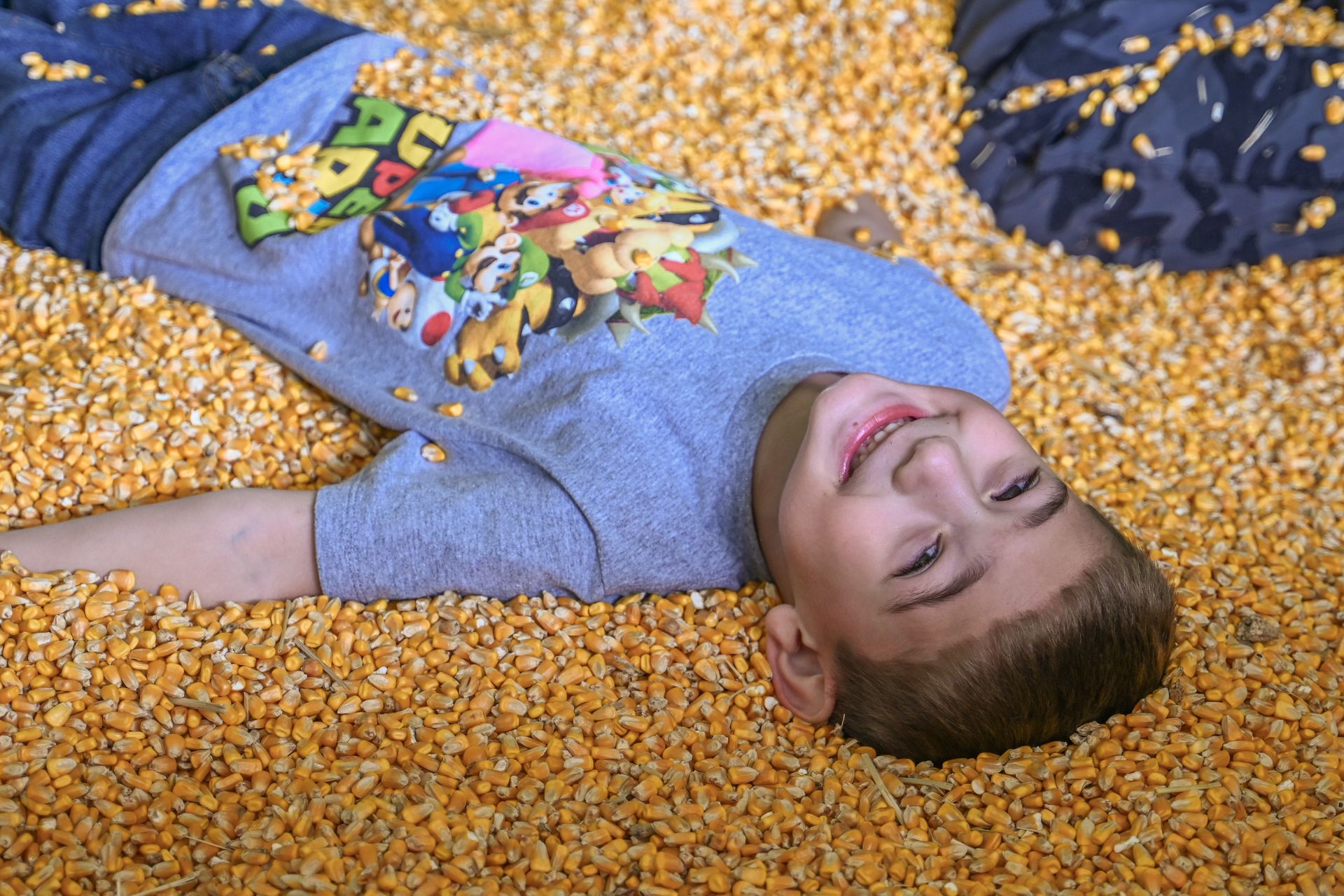 A young boy smiles while lying in a pit of yellow corn kernels.