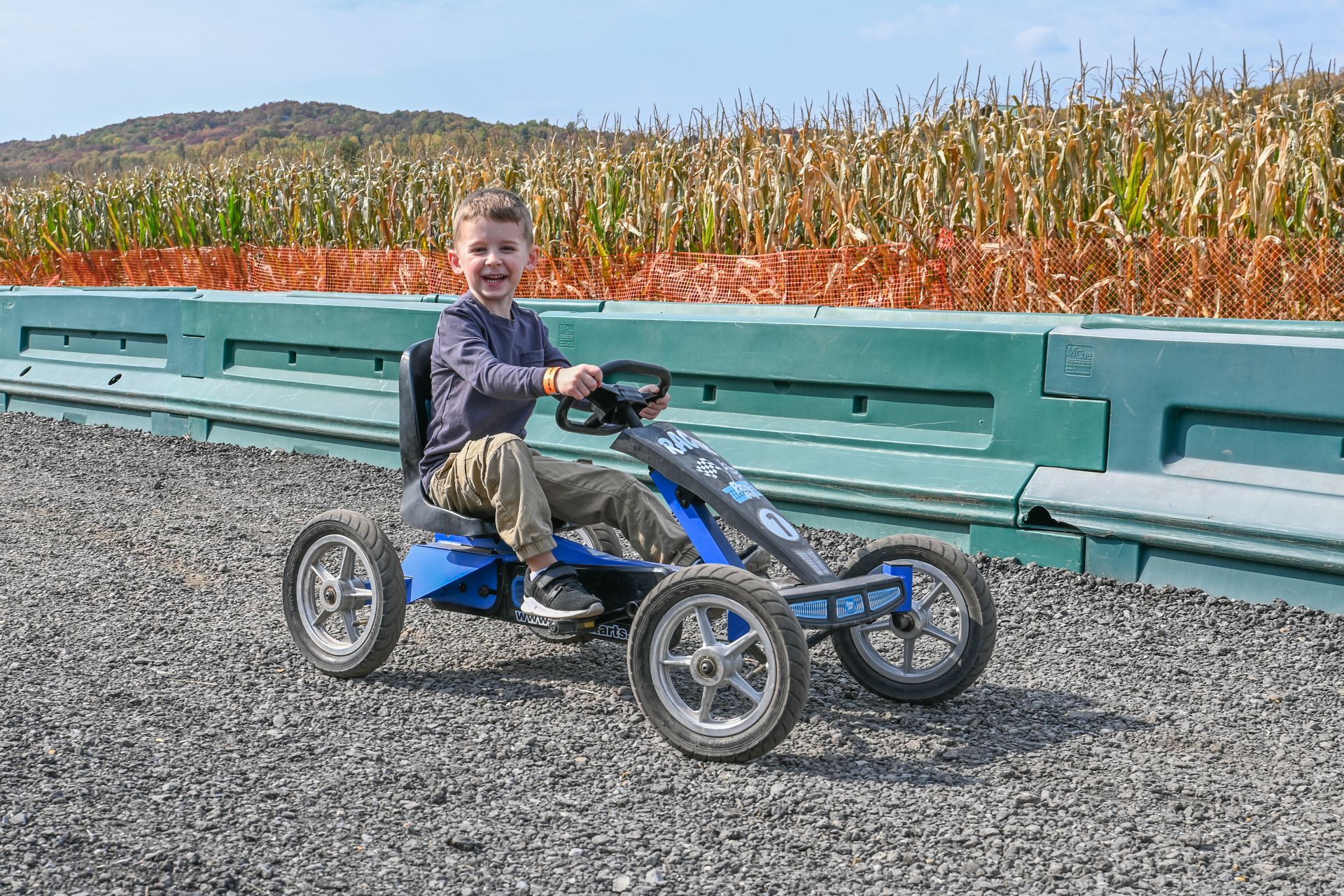 A young boy smiles while driving a blue pedal go-kart on a gravel path, with a cornfield and barriers in the background.