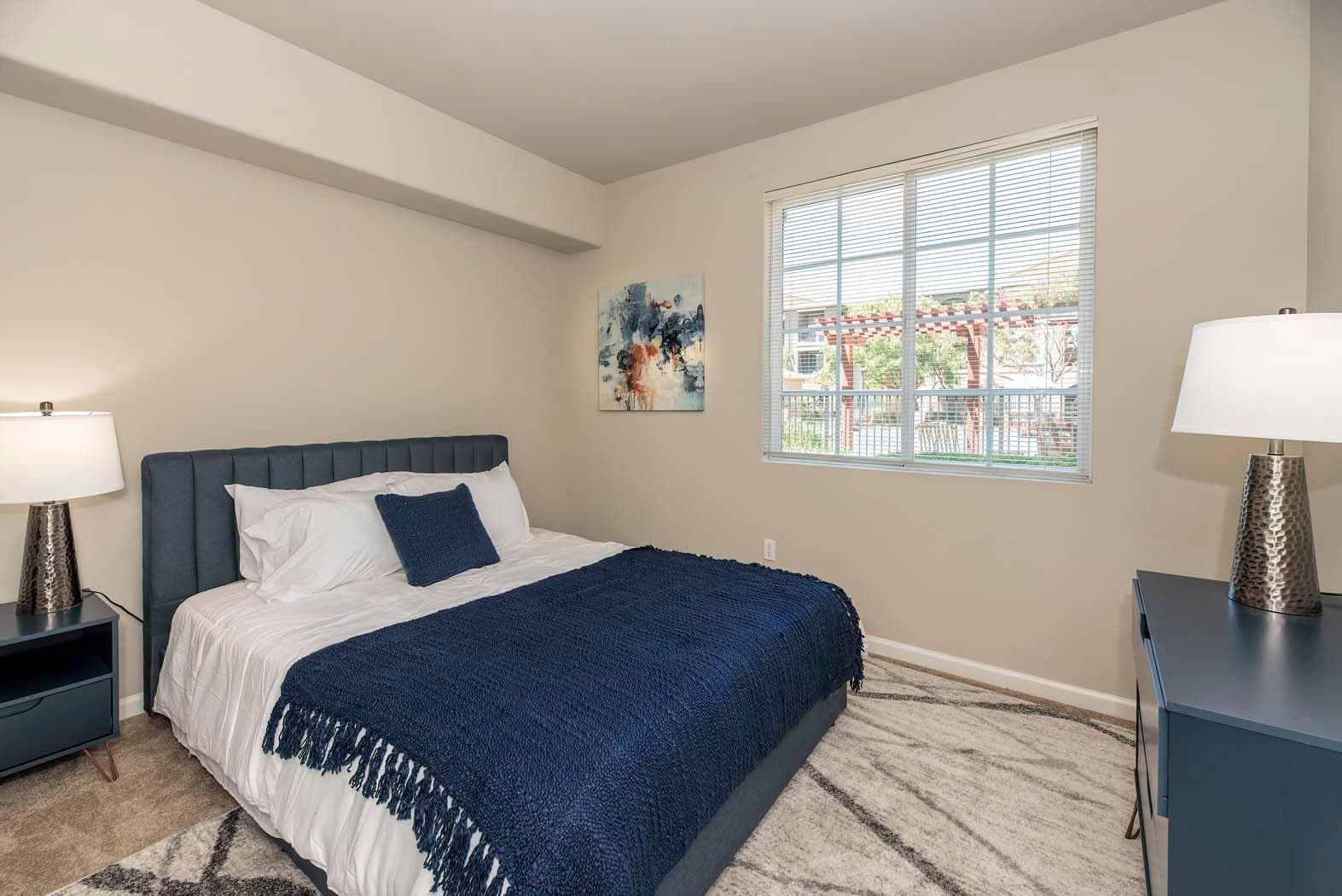 Cozy bedroom with a blue blanket, lamp, and window view.