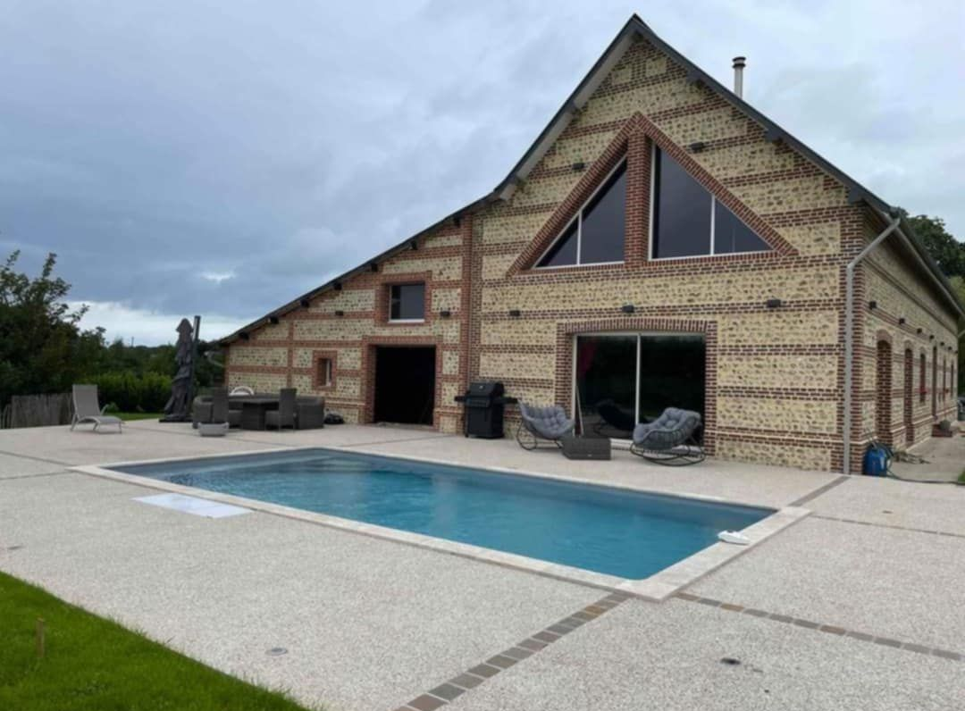 Maison à façade de briques et de bois, fenêtres triangulaires, piscine et ciel nuageux.