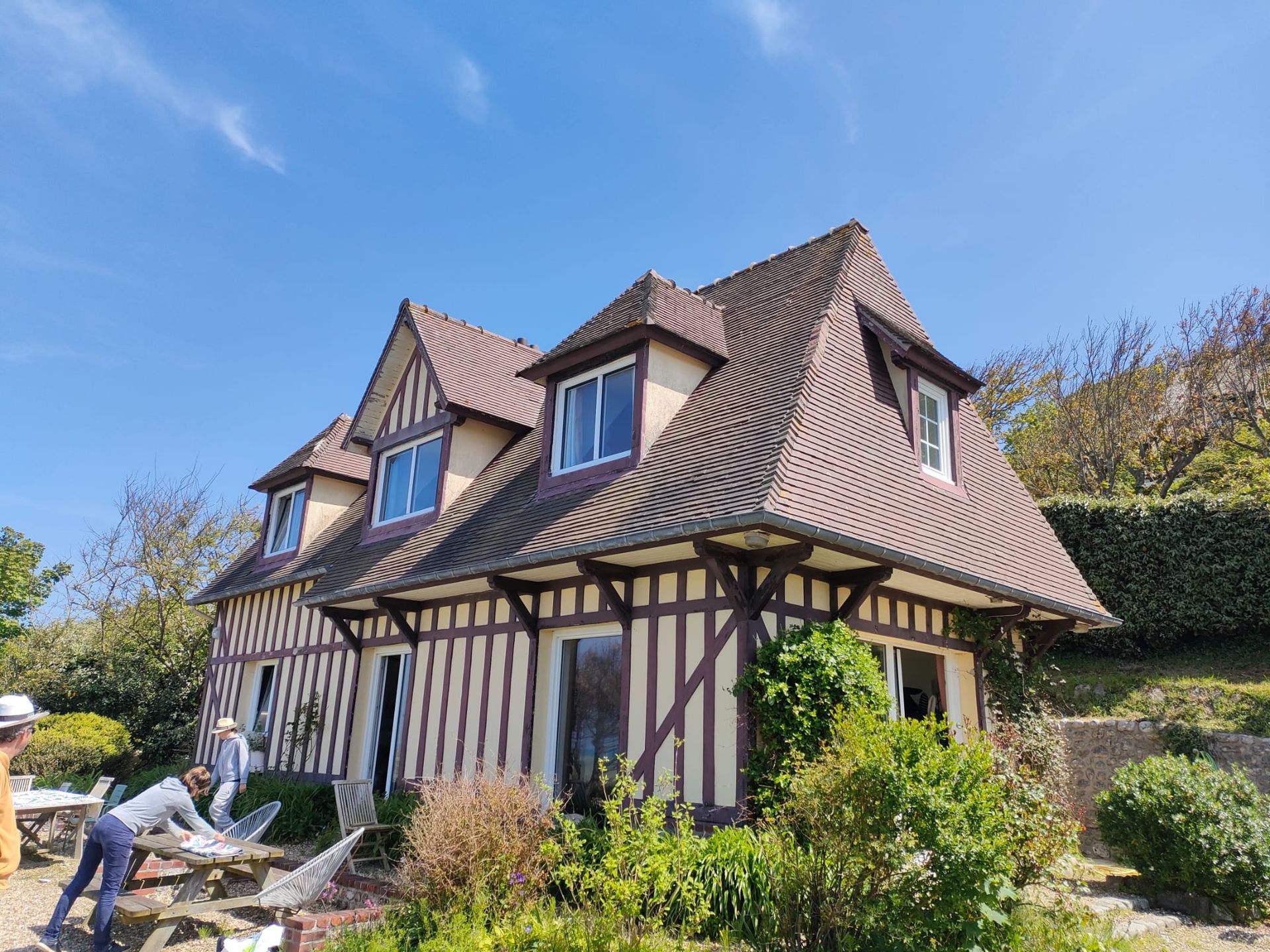 Maison à colombages avec toit et boiseries brunes, murs beiges et trois lucarnes sous un ciel bleu.