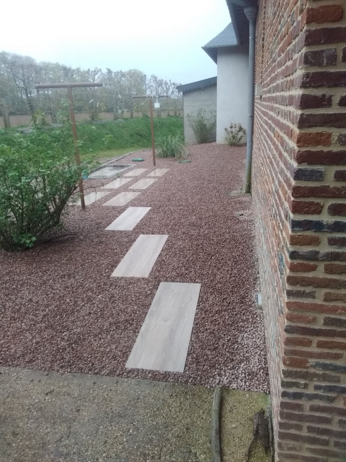 Un chemin de dalles de pierre serpentant à travers du gravier rougeâtre, longeant un bâtiment en briques et un jardin.