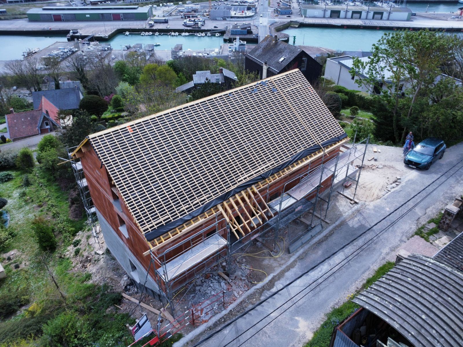 Bâtiment en construction, briques brunes et toit gris. Échafaudages, voiture verte, port en arrière-plan.