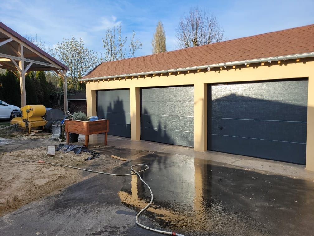 Garage à trois travées avec portes grises, murs beiges et toit rougeâtre. On aperçoit un tuyau d'arrosage et une voiture.