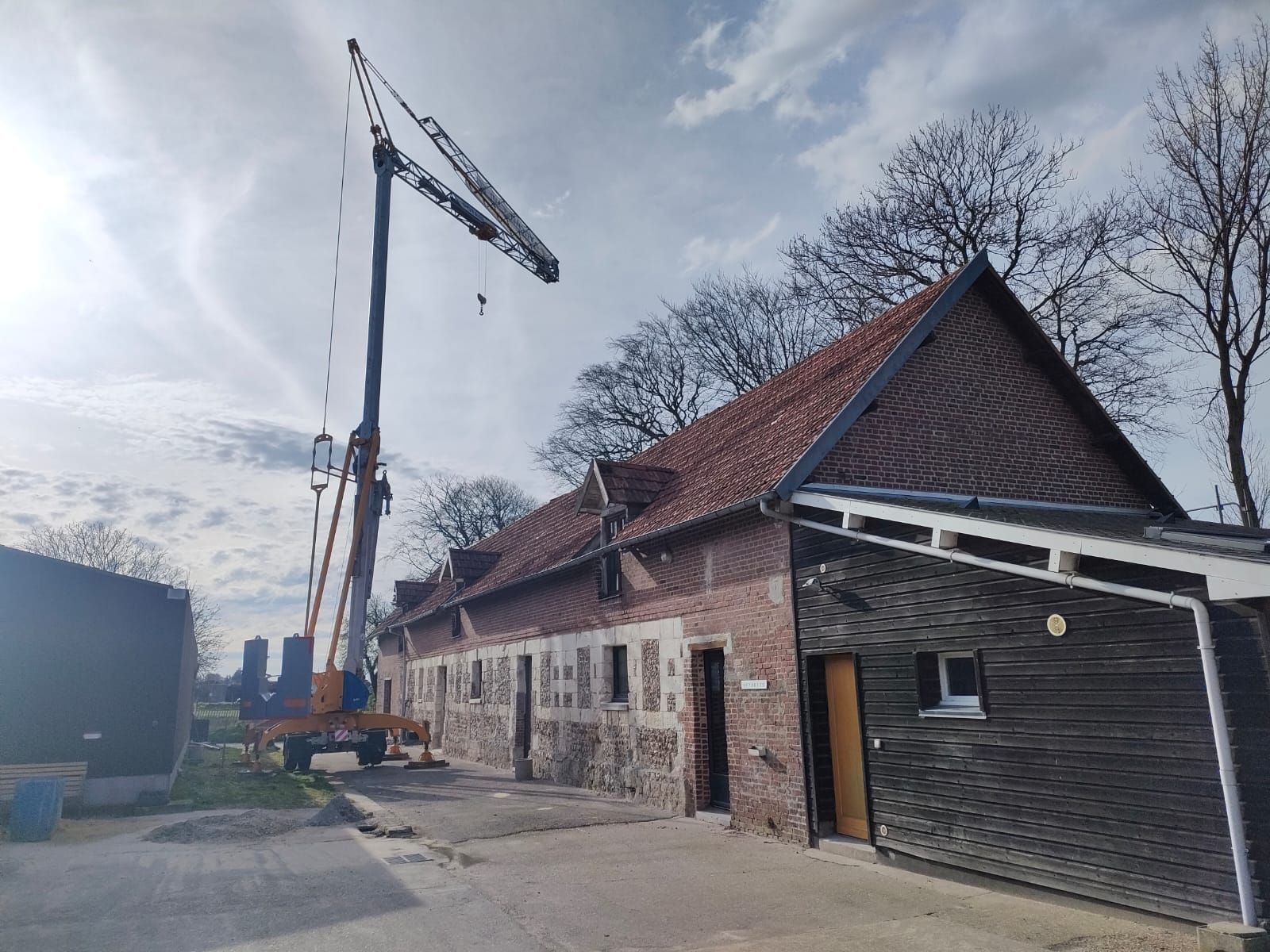Une grande grue près d'un bâtiment en briques au toit de tuiles rouges, sous un ciel nuageux.