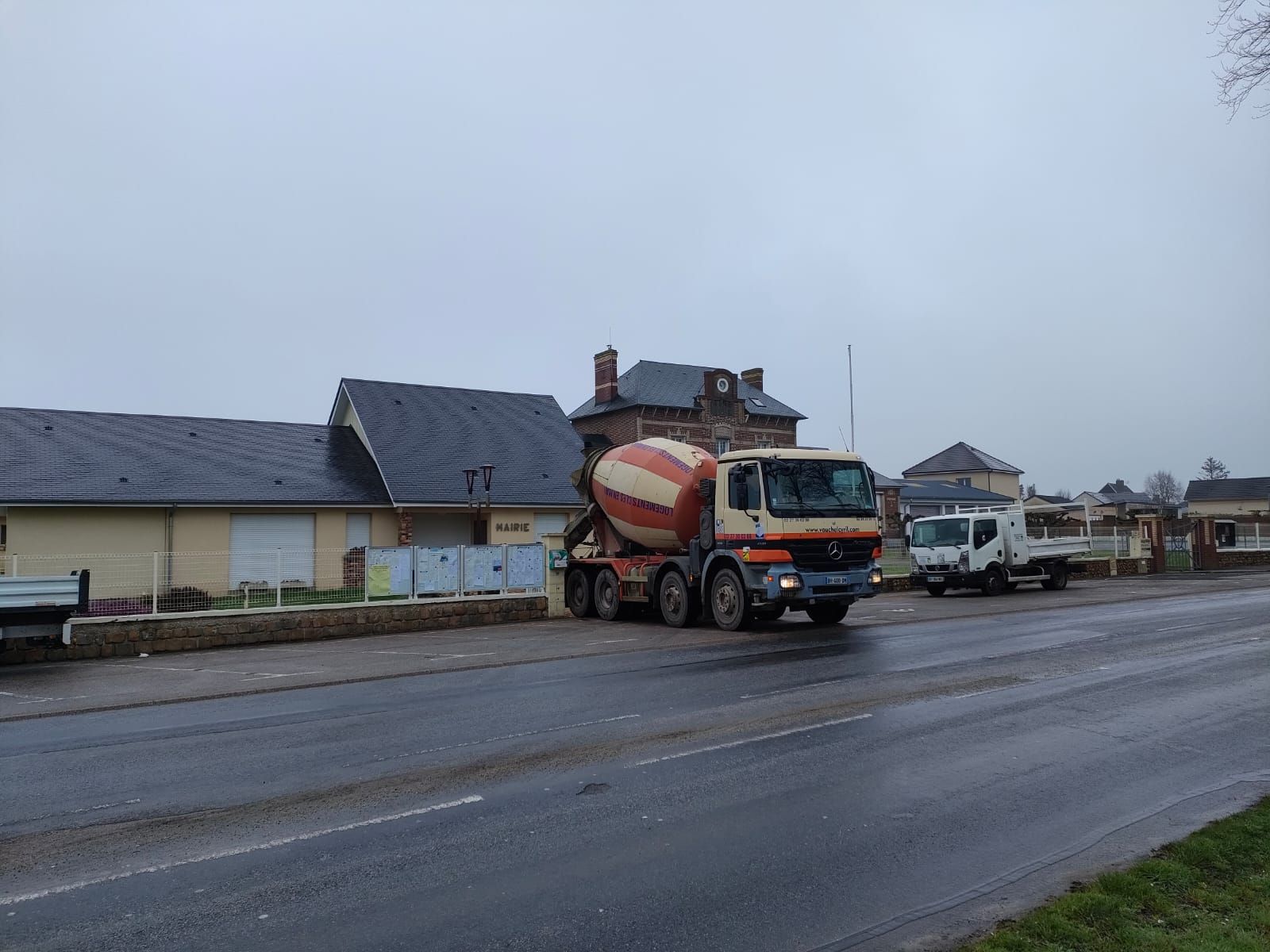 Un camion-citerne à ciment est stationné sur le bord d'une route, devant un bâtiment au toit sombre, par temps couvert.