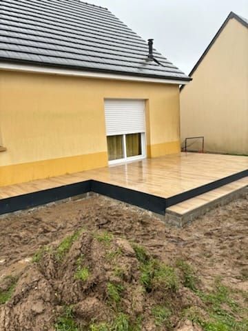 Maison jaune en construction avec terrasse carrelée, donnant sur un terrain boueux.