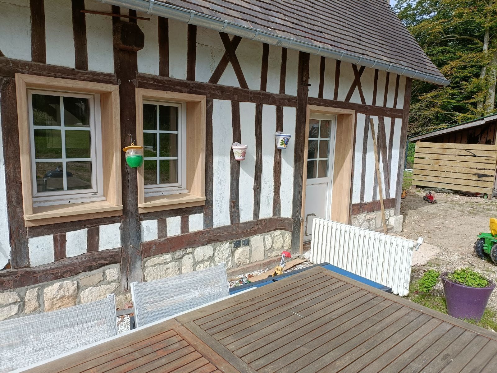 Extérieur d'un bâtiment à colombages avec deux fenêtres, une porte et une terrasse avec une table et des chaises en bois.