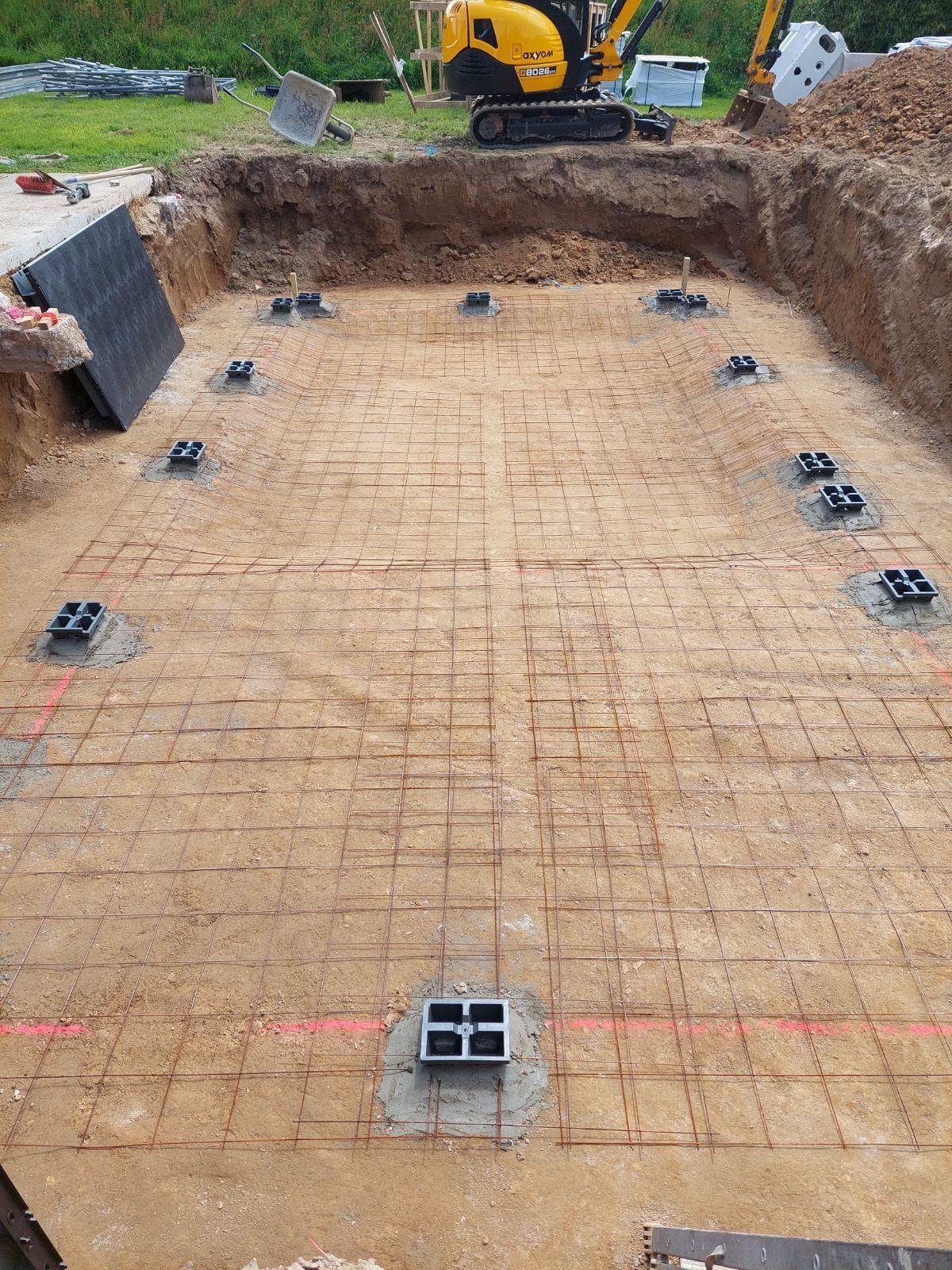 Chantier : fondations rectangulaires excavées, treillis d'armature, supports en béton noir