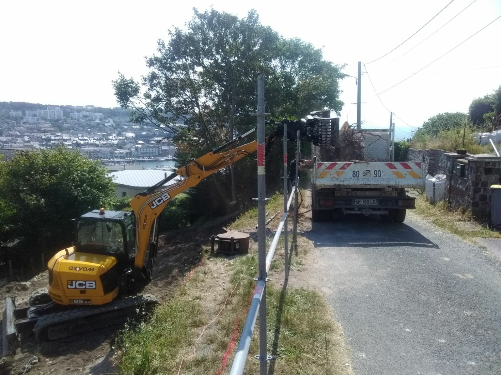 Une pelle mécanique jaune se trouve sur un chemin de terre, à côté d'une clôture et d'un camion stationné