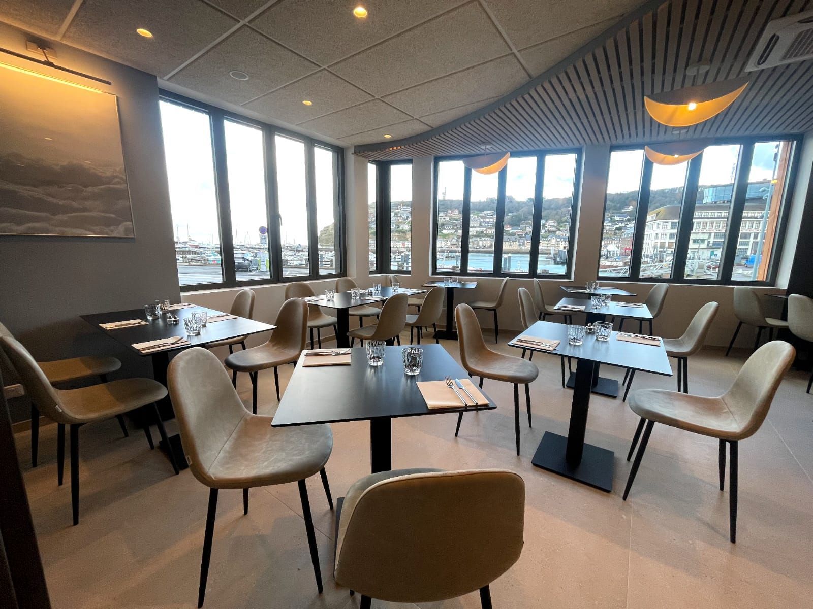 Intérieur de restaurant avec tables, chaises et grandes fenêtres donnant sur le port. Décoration beige et noire.