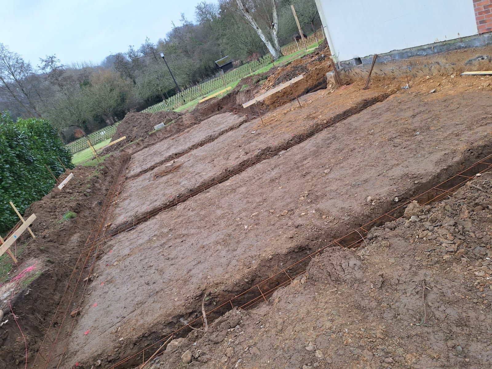 Tranchées de fondation creusées dans une cour, avec de la terre et des planches de bois, près d'un bâtiment.