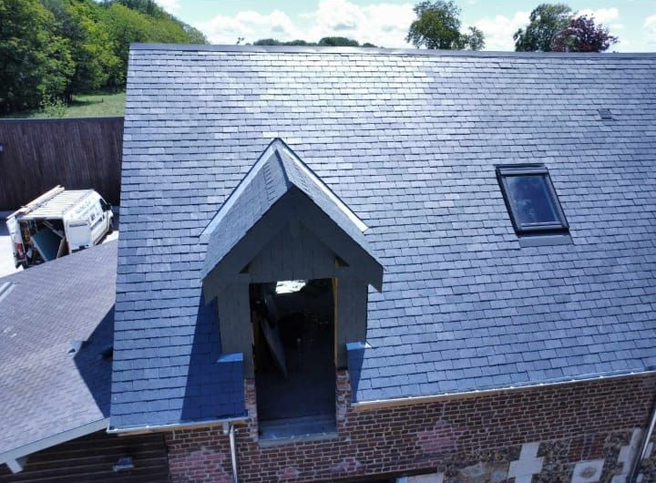 Toit en ardoise grise avec lucarne, puits de lumière et façade en briques.