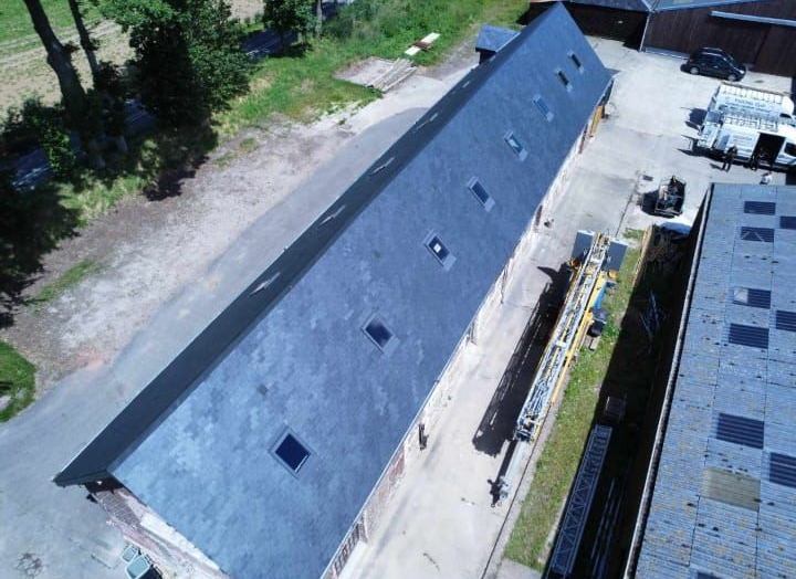 Vue aérienne d'un long bâtiment au toit sombre percé de plusieurs puits de lumière, entouré d'une zone industrielle