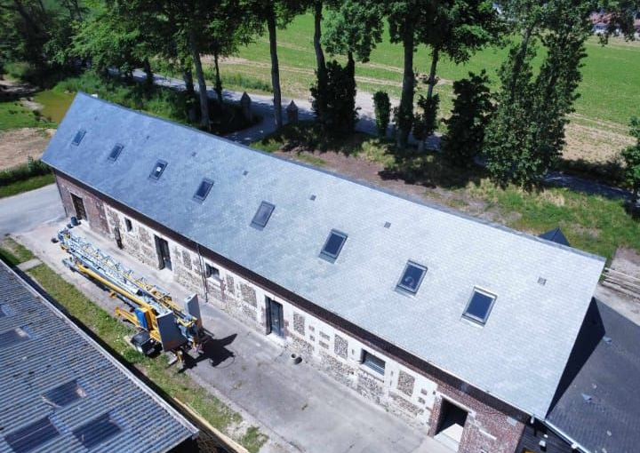 Long bâtiment en briques, toit d'ardoise grise, plusieurs puits de lumière et volets fermés.