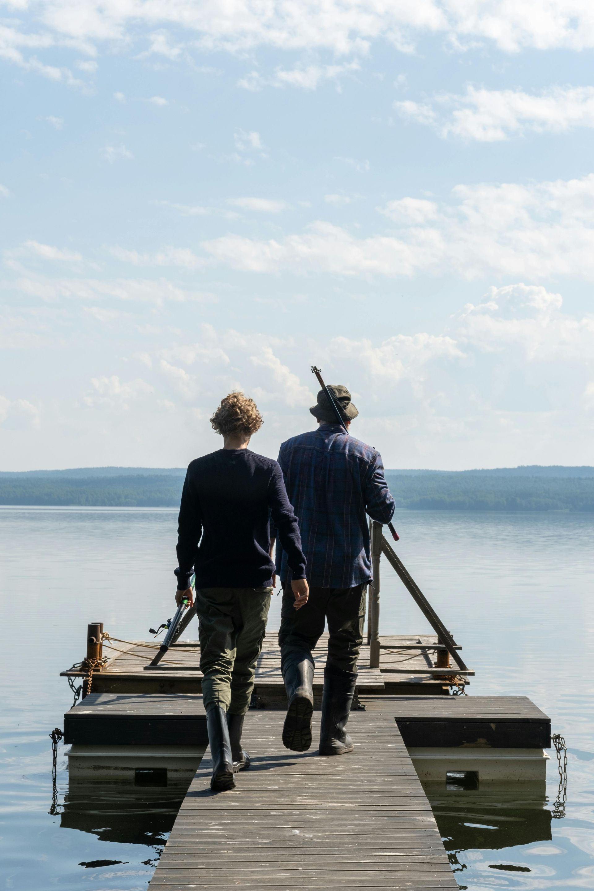 Deux personnes marchent sur un quai en bois en direction d'un lac calme sous un ciel nuageux.