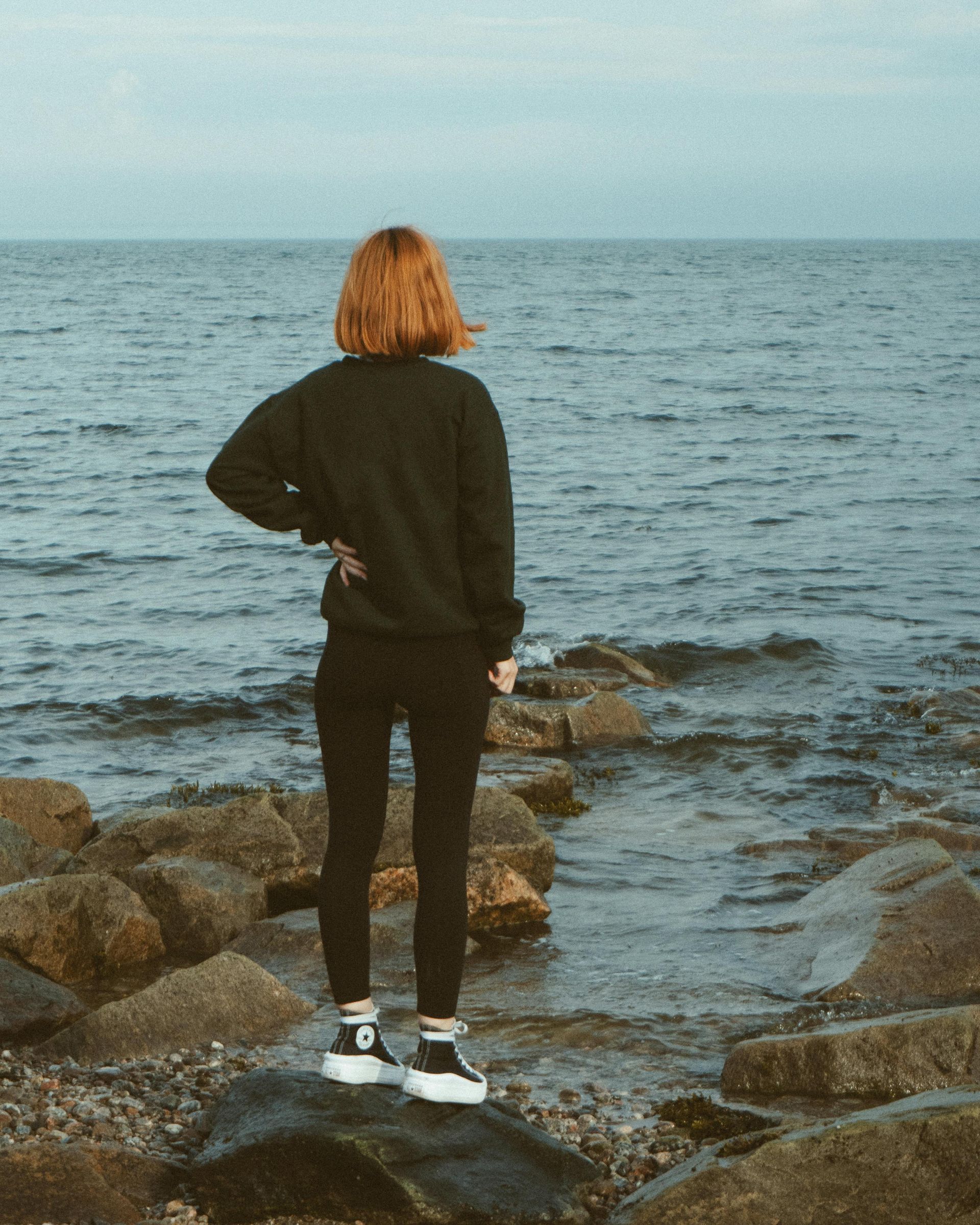 Une personne se tient debout sur des rochers, contemplant l'océan. Elle porte un sweat-shirt noir