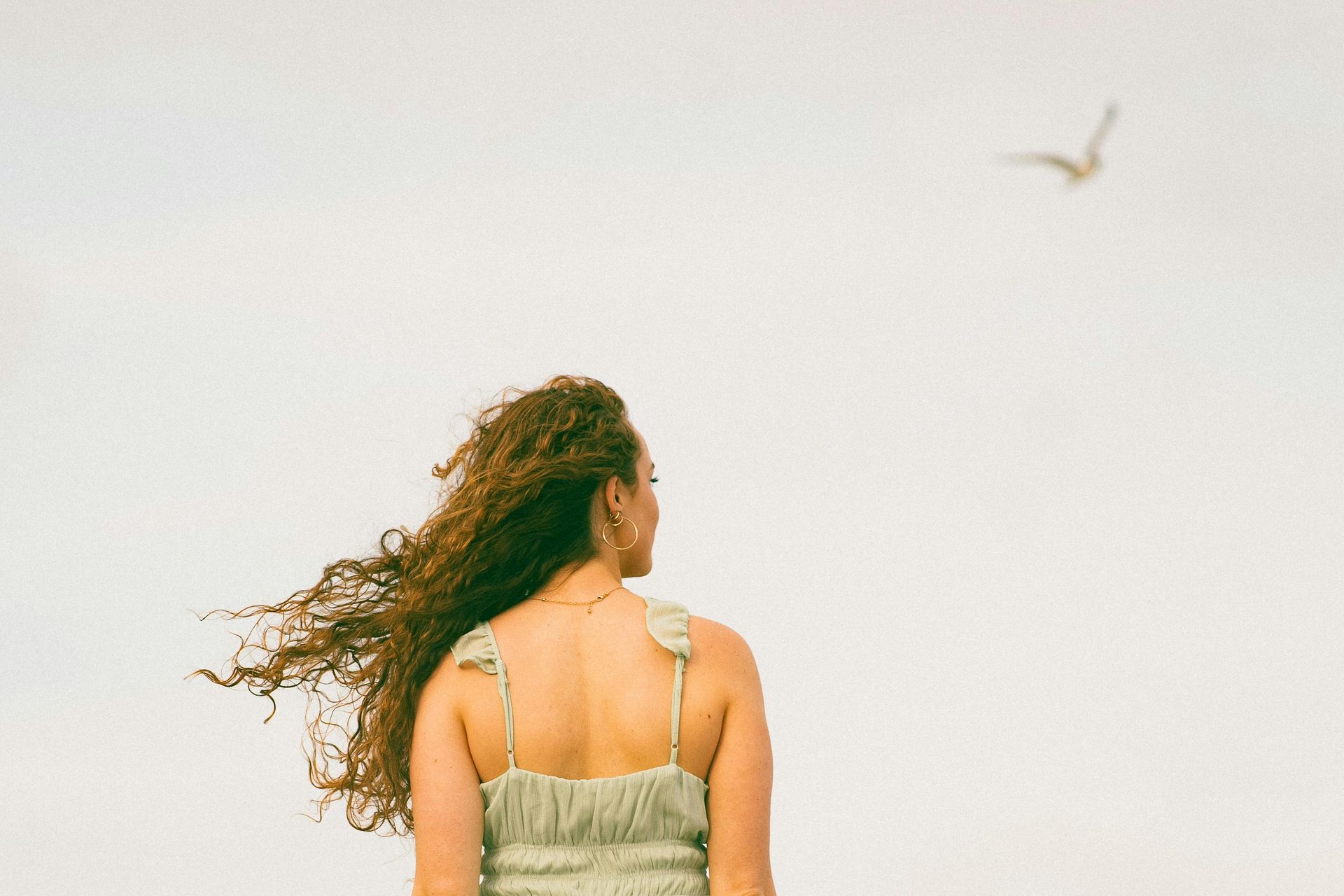 Femme aux cheveux bouclés regardant une mouette dans un ciel clair ; vêtue d'une robe vert clair.