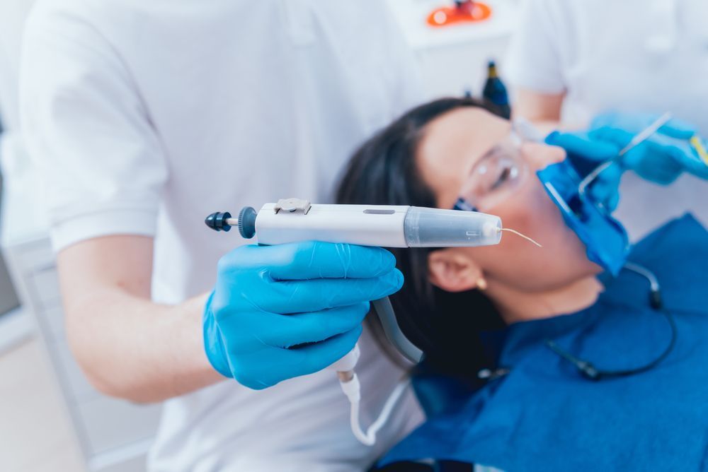 A dental professional in blue gloves holds a digital dental injection device near a patient in a dental chair.