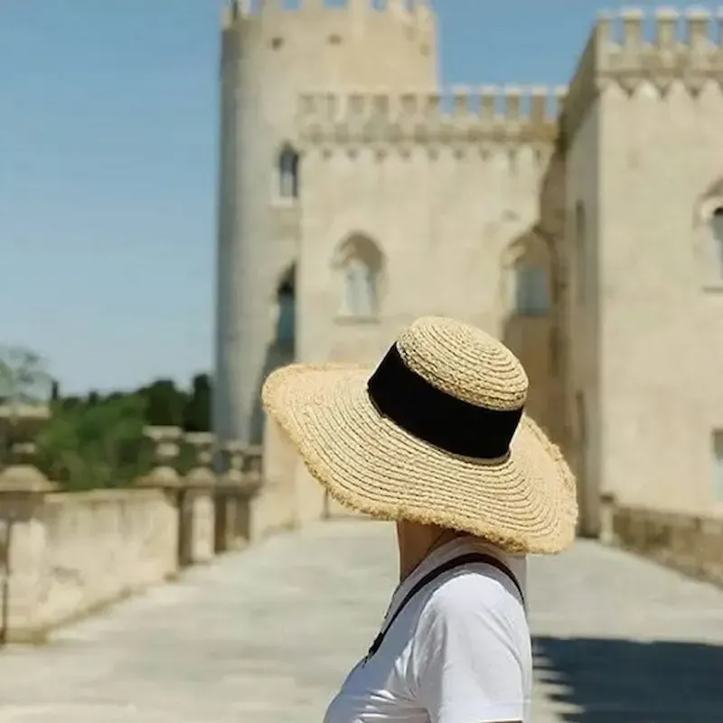 Person in a straw hat walking near a stone castle under a clear blue sky