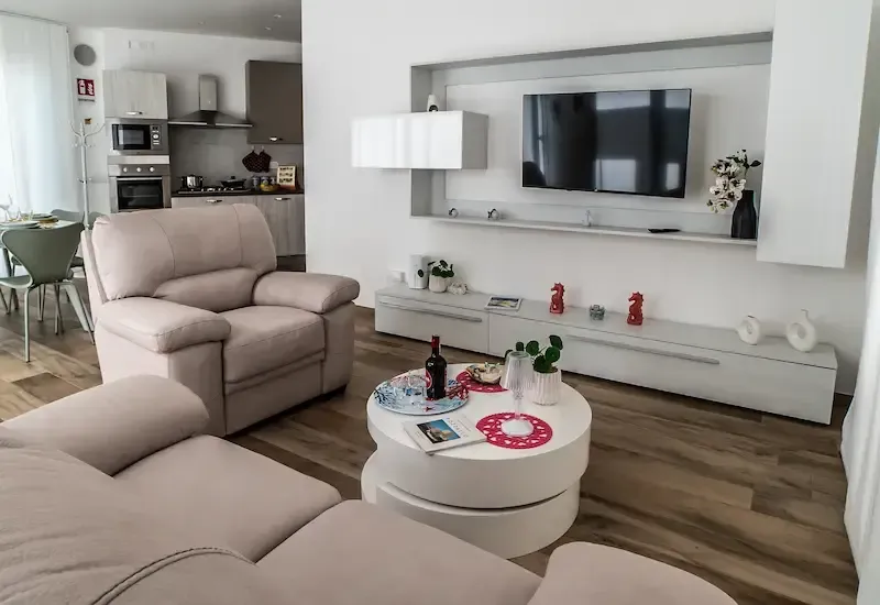 Bright modern living room with beige sofa, wall-mounted TV, white coffee table, and open kitchen in the background