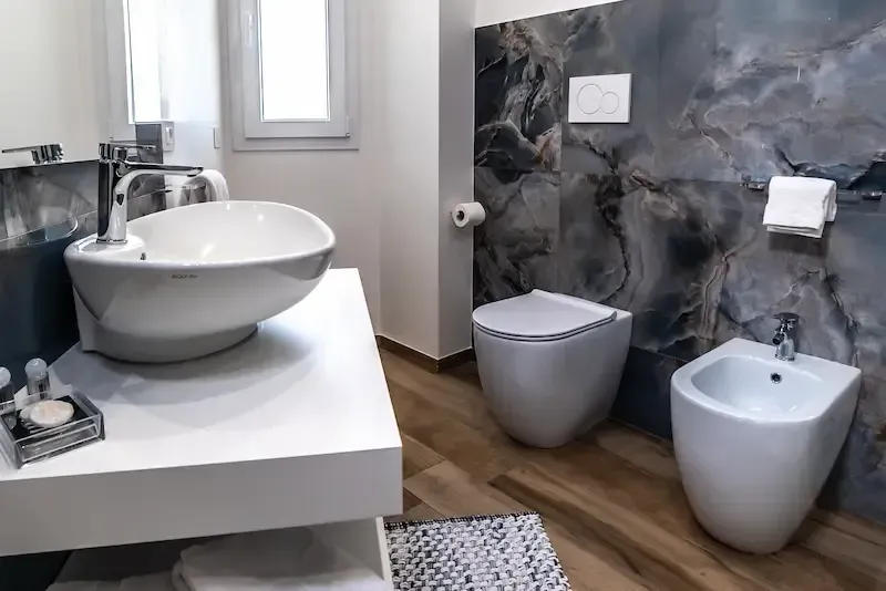 Modern bathroom with white sink, gray bidet and toilet, dark stone wall, and wood floor