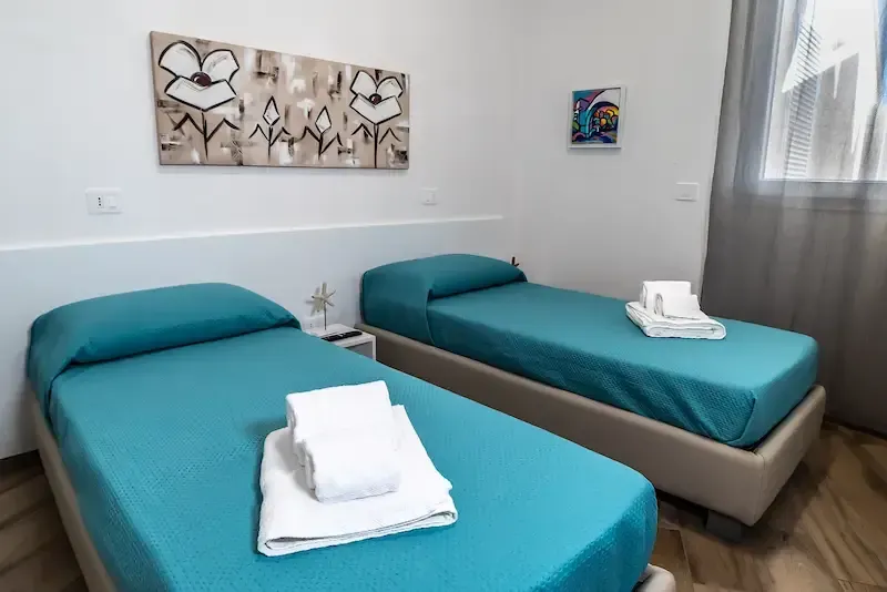 Bright twin room with two teal beds, folded towels, and wall art in a white interior