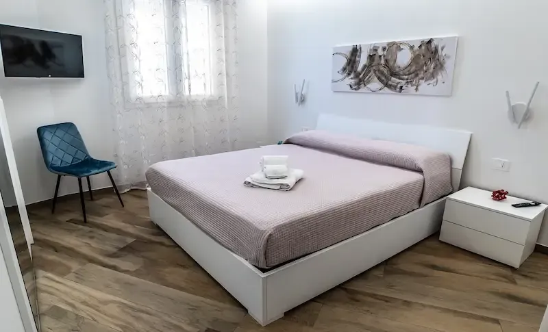 Minimalist white bedroom with a double bed, blue chair, wall art, and a small side table on wood floors