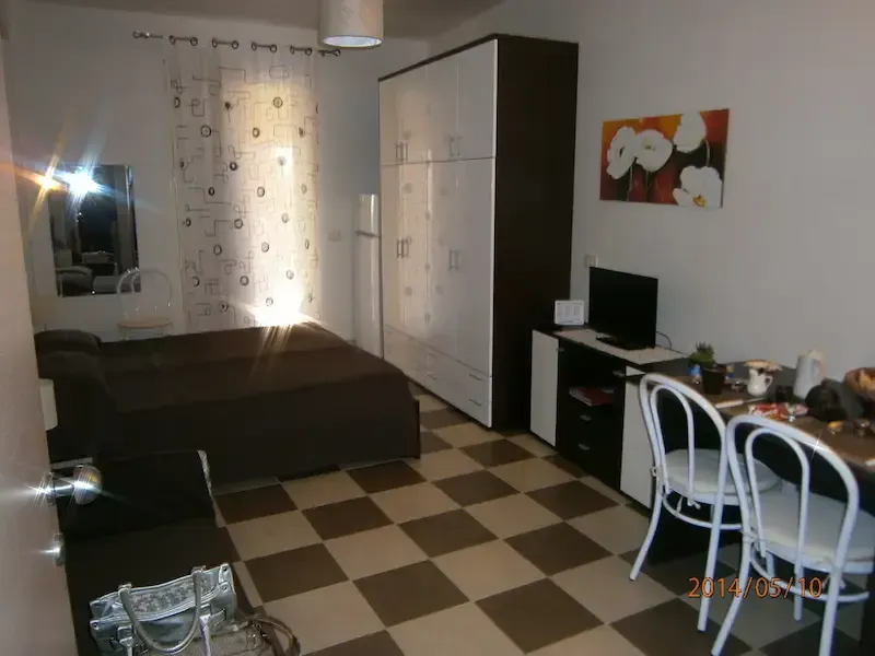Small room with checkerboard floor, bed, wardrobe, desk, and chairs near a window with curtains.