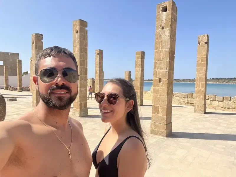 Two visitors standing among ancient stone columns near the sea in Vendicari.